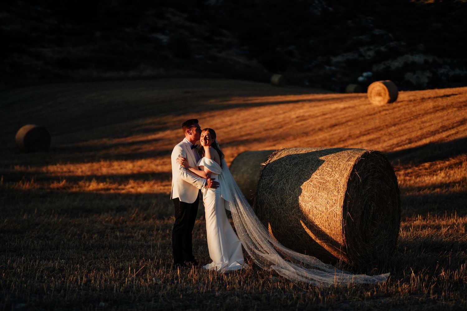 Bride and groom embracing at golden hour beside hay bales in a sunlit field, bride wearing a flowing white gown with long veil.