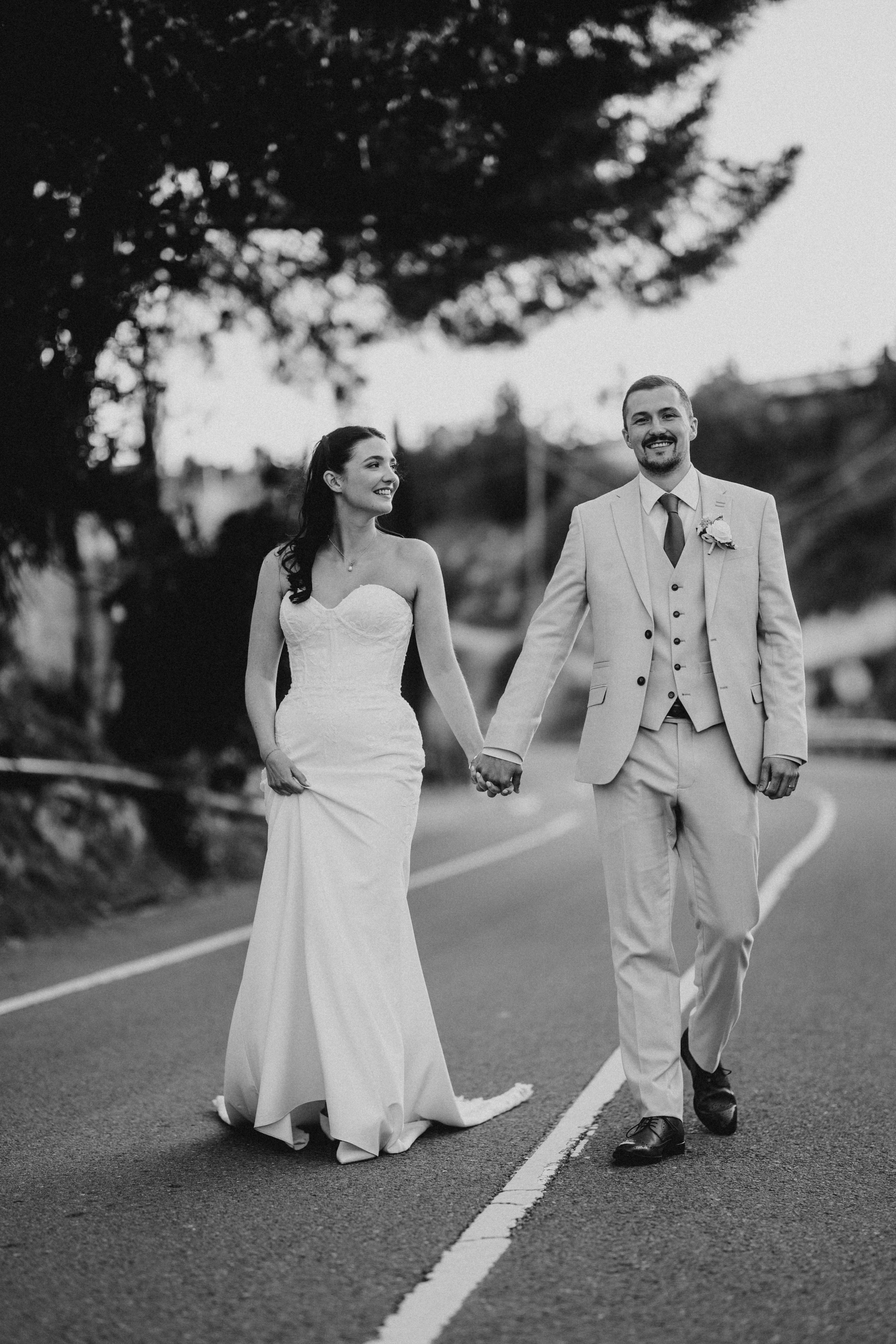 Black and white photo of a smiling bride and groom holding hands walking down a road on their wedding day.