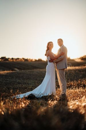 Bride and groom embracing in golden field at sunset, bride in white gown with train, groom in tan suit.