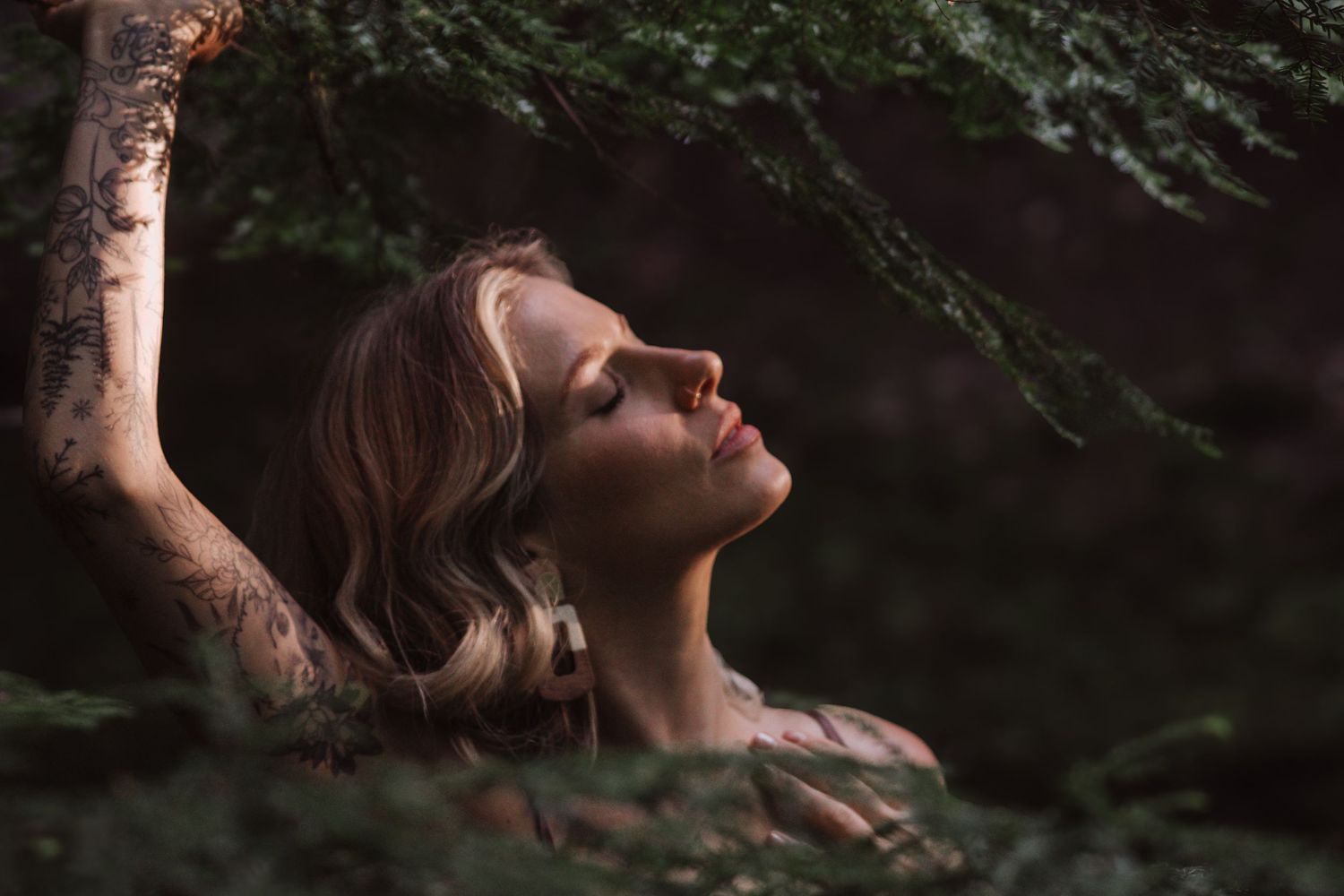 Woman with tattoos tilts head back among green forest branches in moody nature portrait.