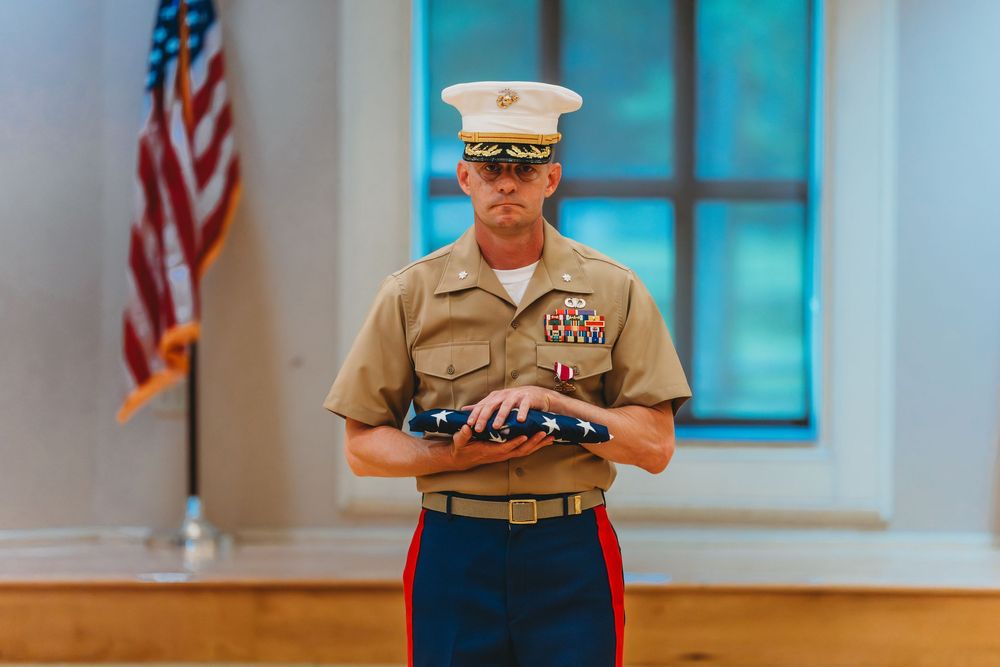 A US Marine in dress uniform holds a folded American flag during a formal military ceremony indoors.
