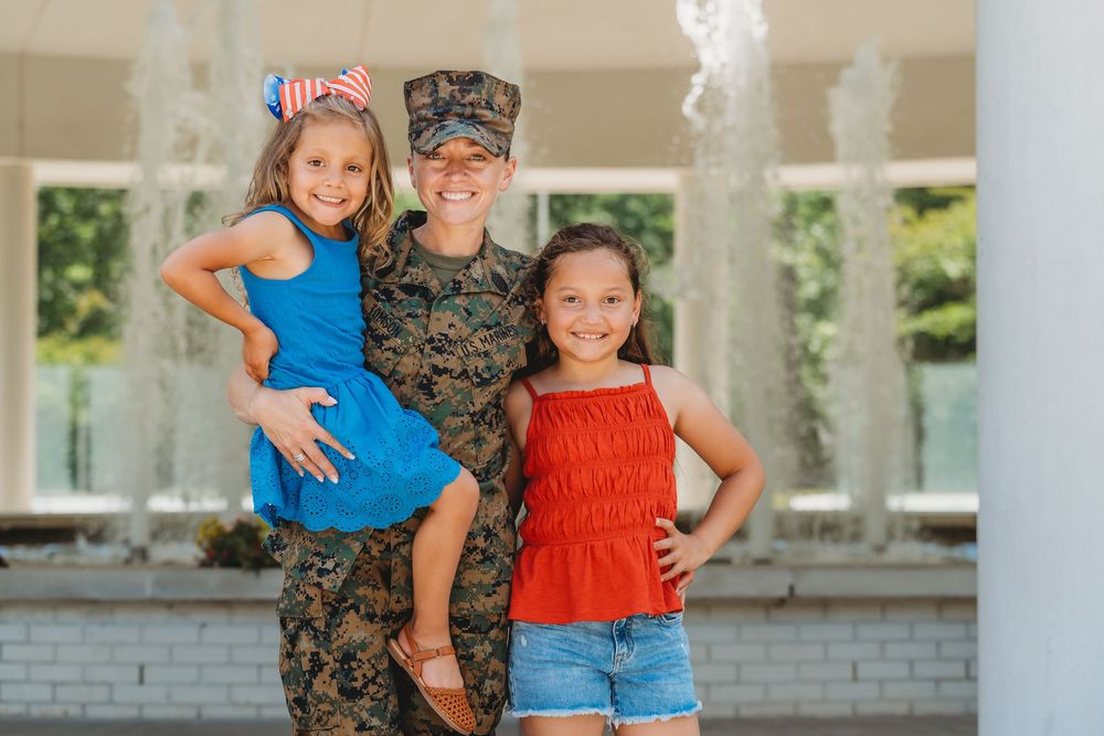 Female soldier in camouflage uniform smiling with two young girls in front of a fountain.