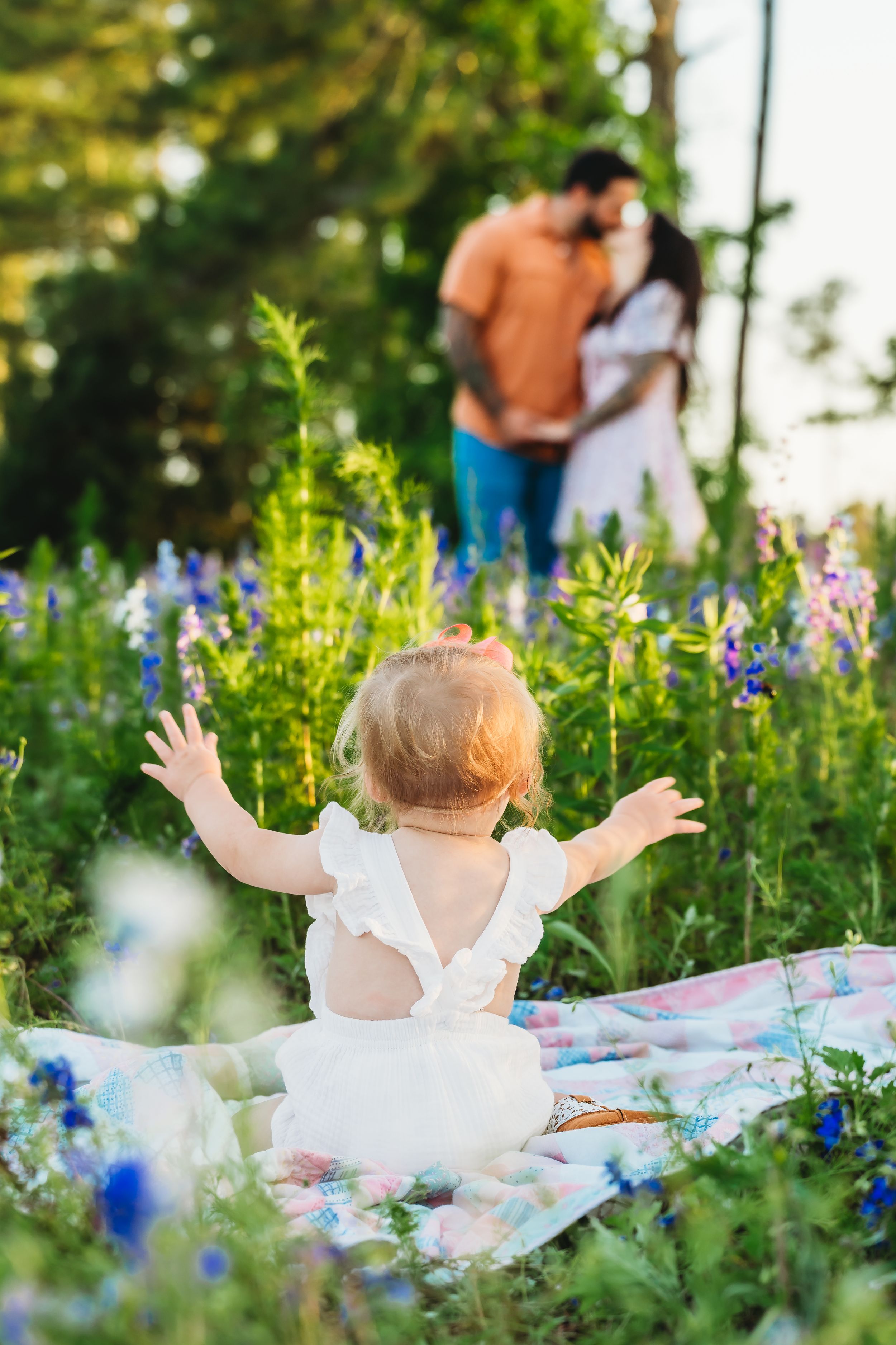Baby in white dress sits on blanket in flower field, arms raised toward couple embracing in sunlit meadow background.