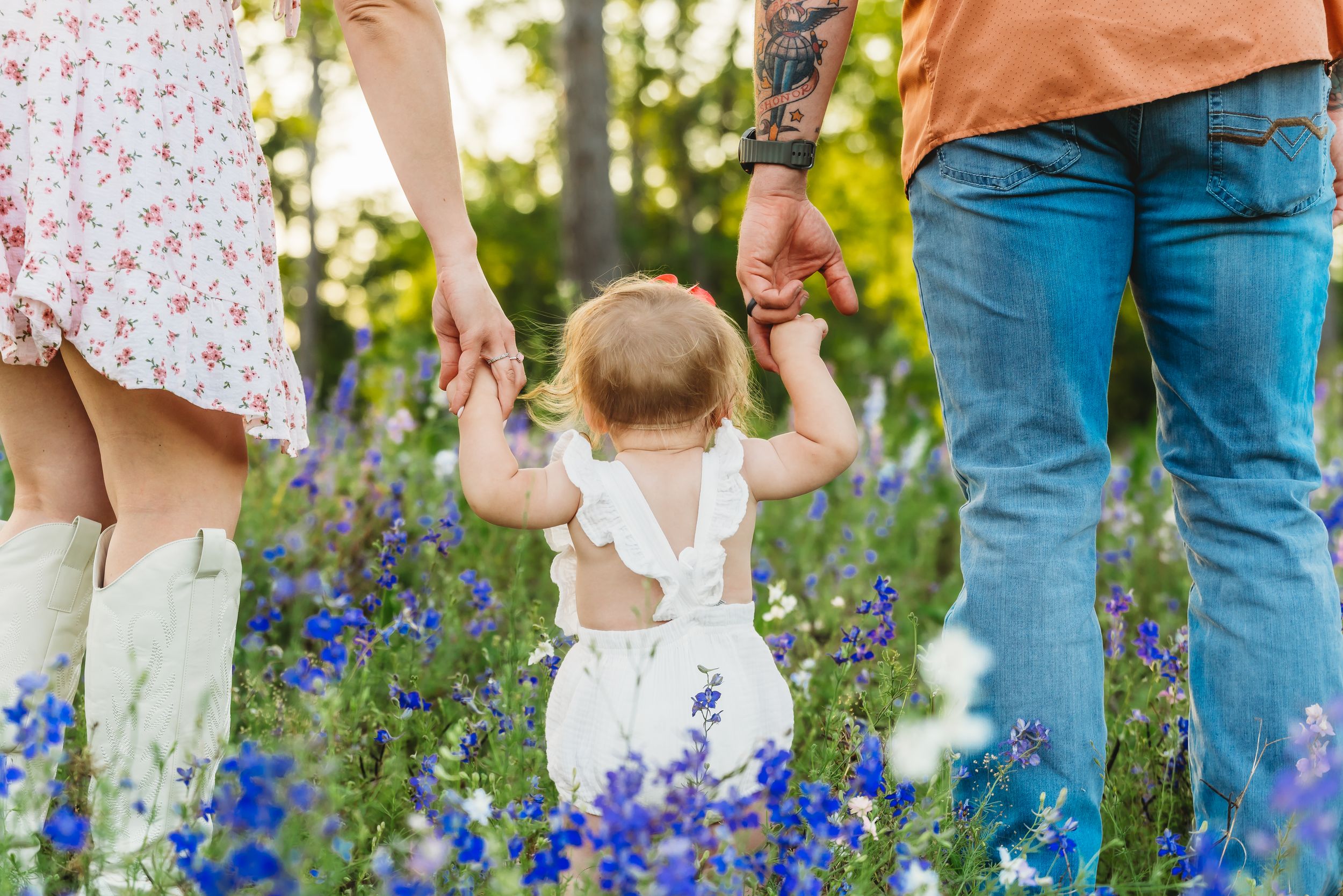 A baby takes first steps holding parents hands in a field of blue wildflowers during a family photo session.