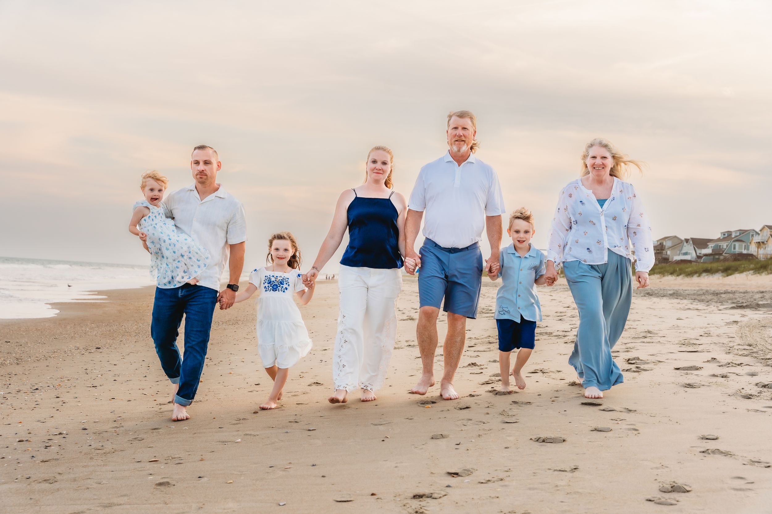 Multi-generational family walking on beach at sunset, wearing coordinated blue and white outfits, smiling together.