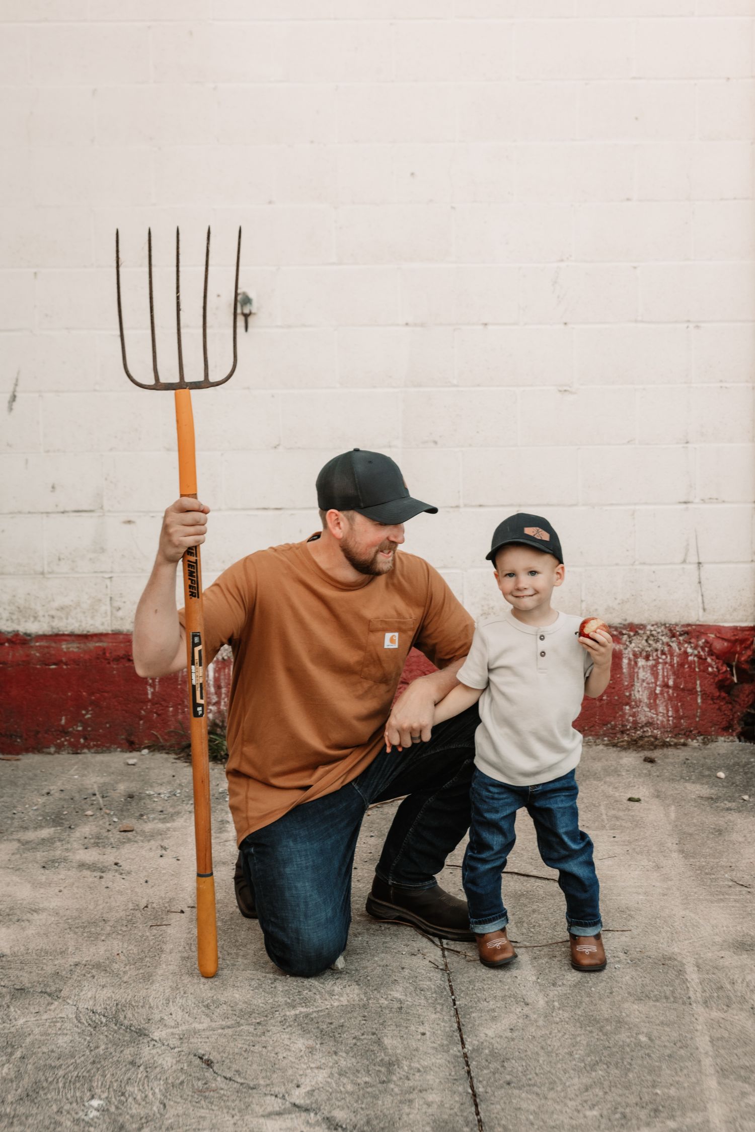Two people pose together against a white wall, with one holding a large pitchfork while kneeling on concrete.