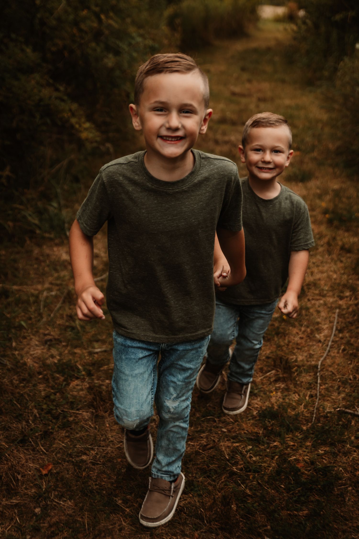 Two children in matching gray shirts run and play on a wooded trail.