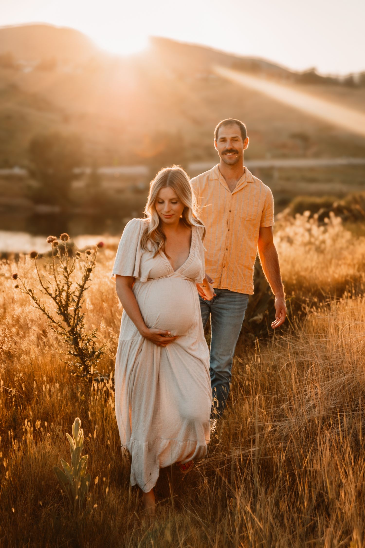 A couple stands in golden sunlit field during sunset maternity photoshoot.