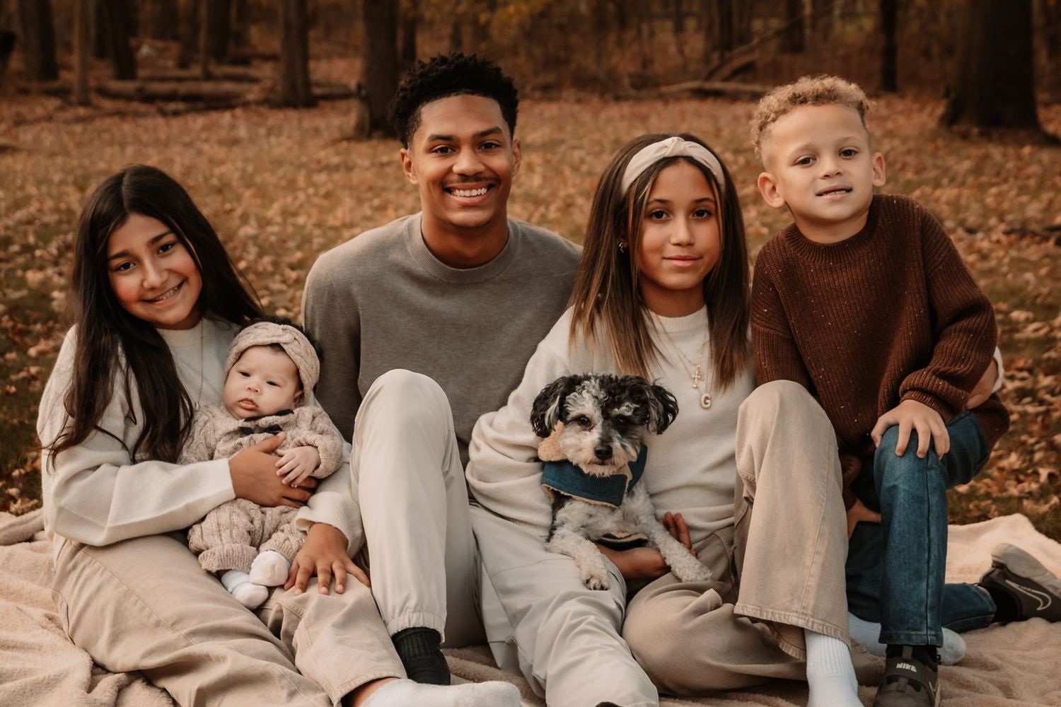 Family sits together on autumn ground with their dog and children in coordinated neutral outfits.