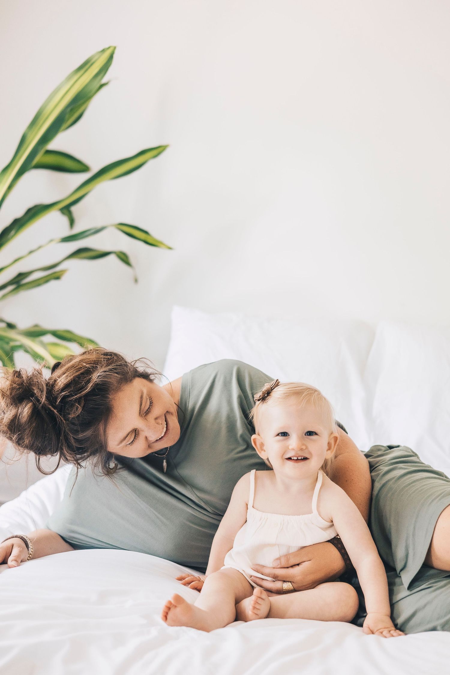 Mother smiling at happy baby sitting on white bed with green plant background.