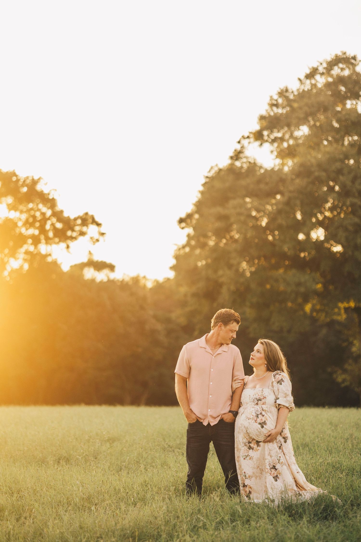 Couple standing in a sunlit field at golden hour, woman in floral dress holding baby bump, man in pink shirt smiling at her.