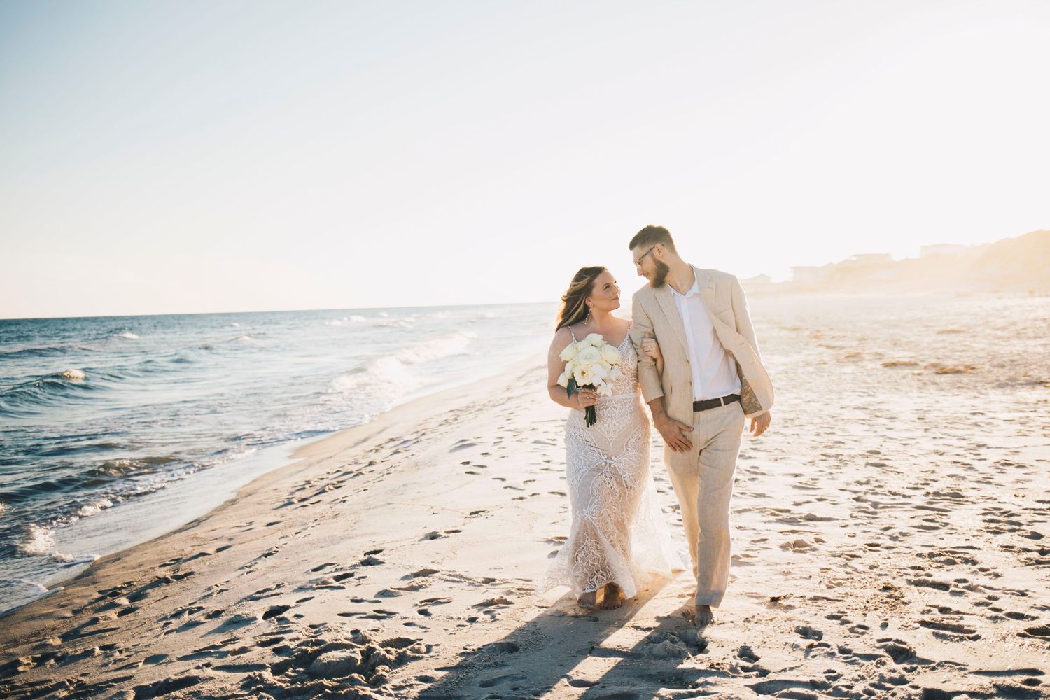 Newlywed couple walking hand-in-hand along a sunlit beach after their wedding ceremony.