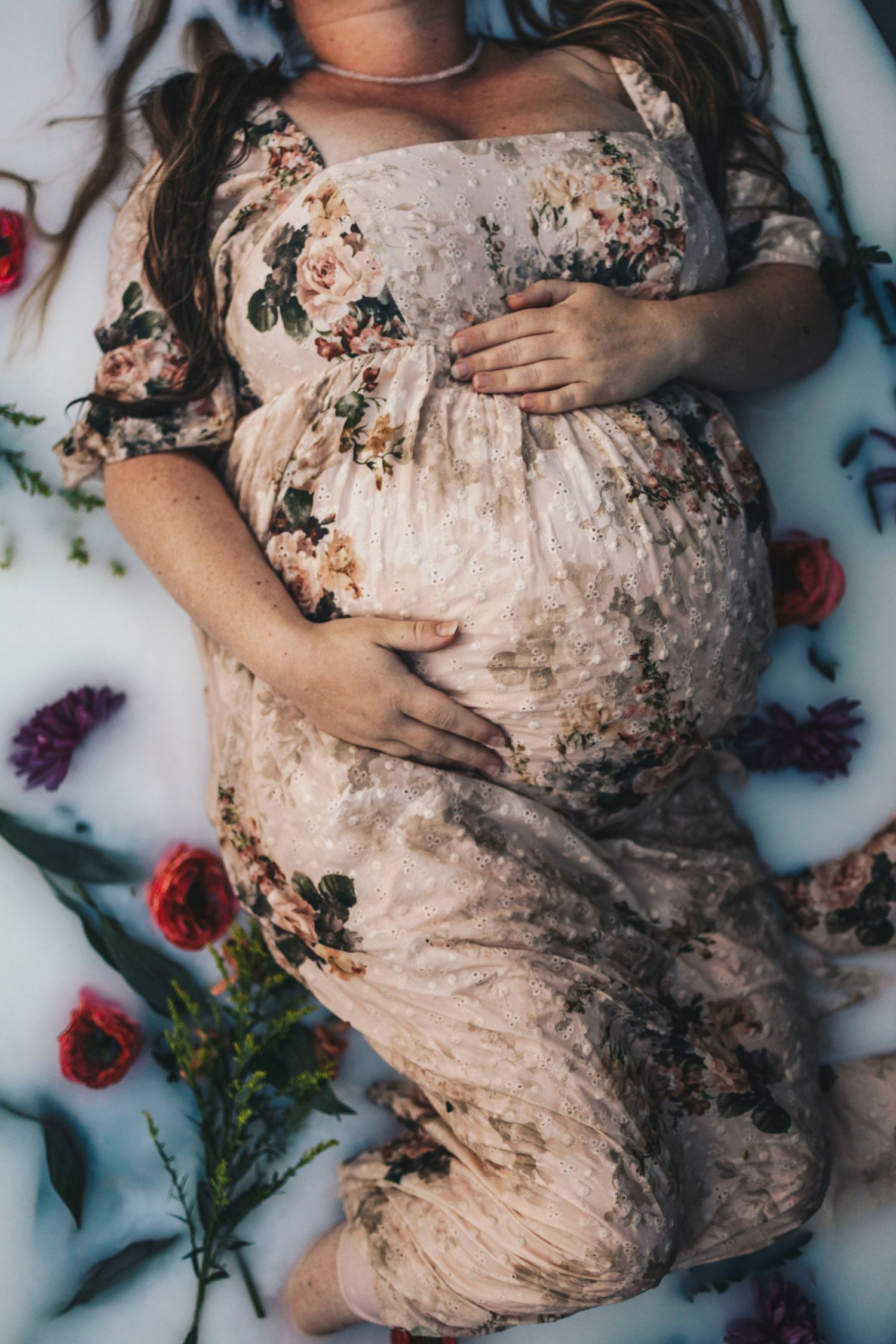 Pregnant woman in floral dress lying in milk bath surrounded by colorful flowers, cradling her baby bump.