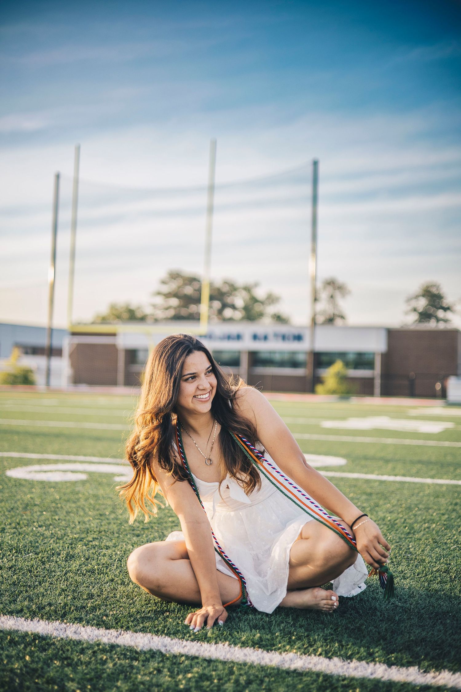 Smiling young woman in white dress with graduation sash sitting on green football field turf.