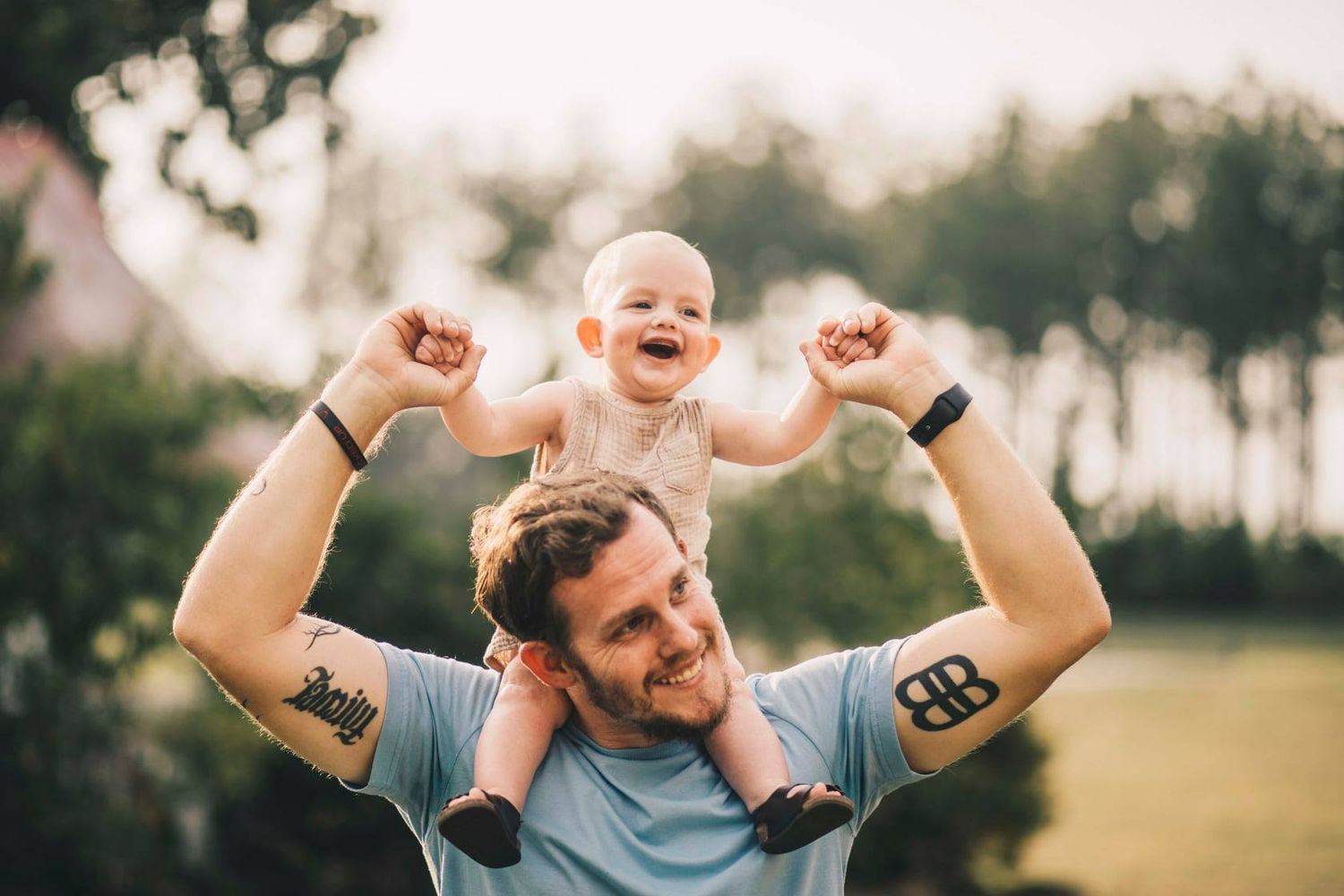 Happy baby sitting on fathers shoulders outdoors, laughing with arms raised in a sunny green park.