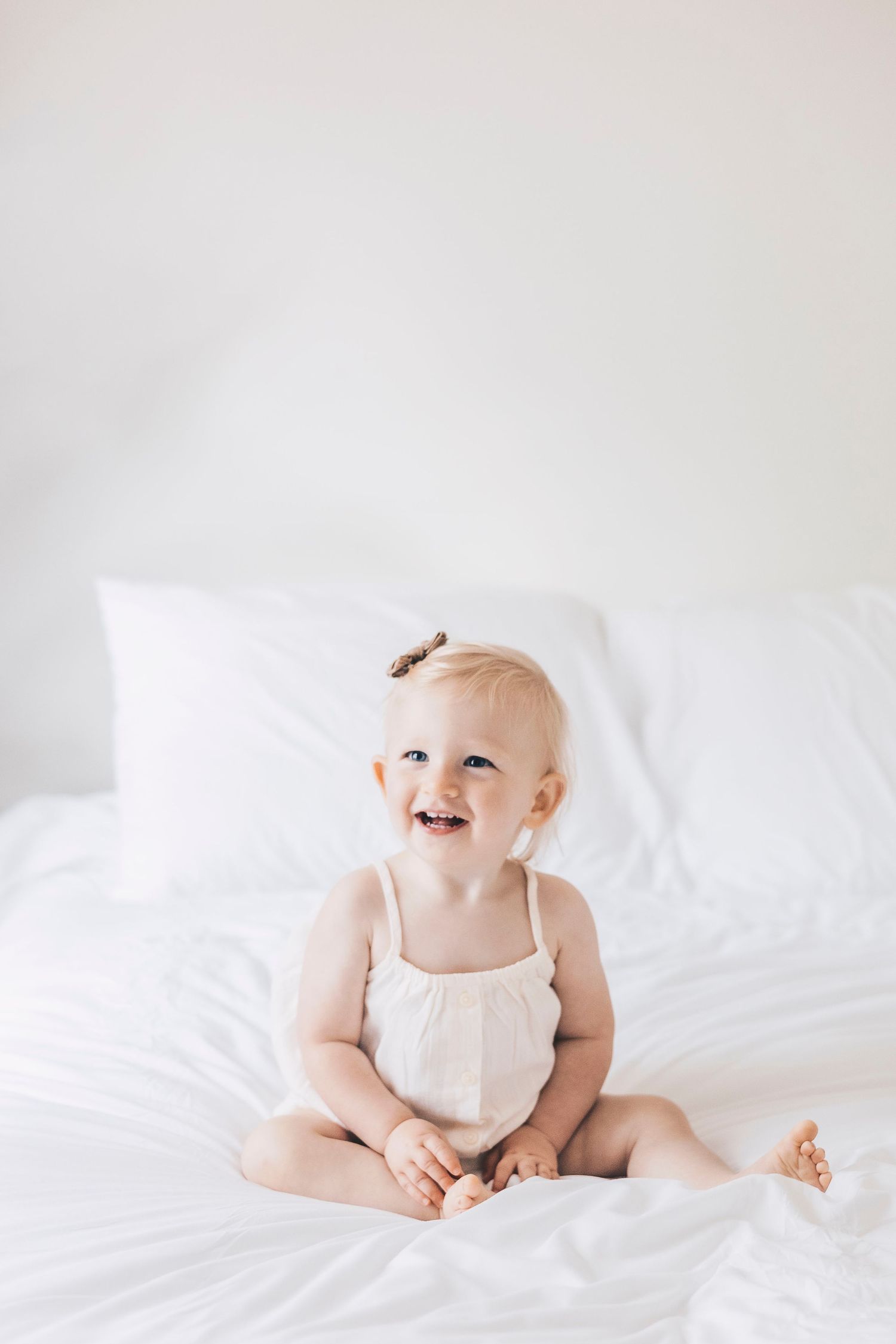 Smiling baby girl in white romper sitting on white bed looking up.