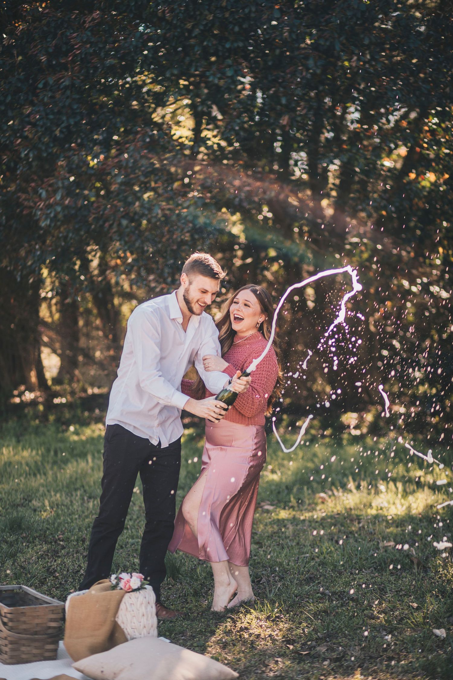 A joyful couple laughs as champagne sprays during an outdoor picnic celebration.