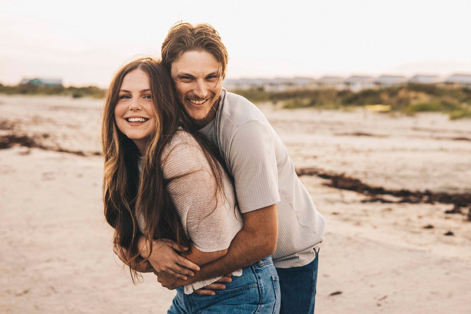 A happy couple embraces on a sandy beach, smiling joyfully in casual clothes during golden hour.