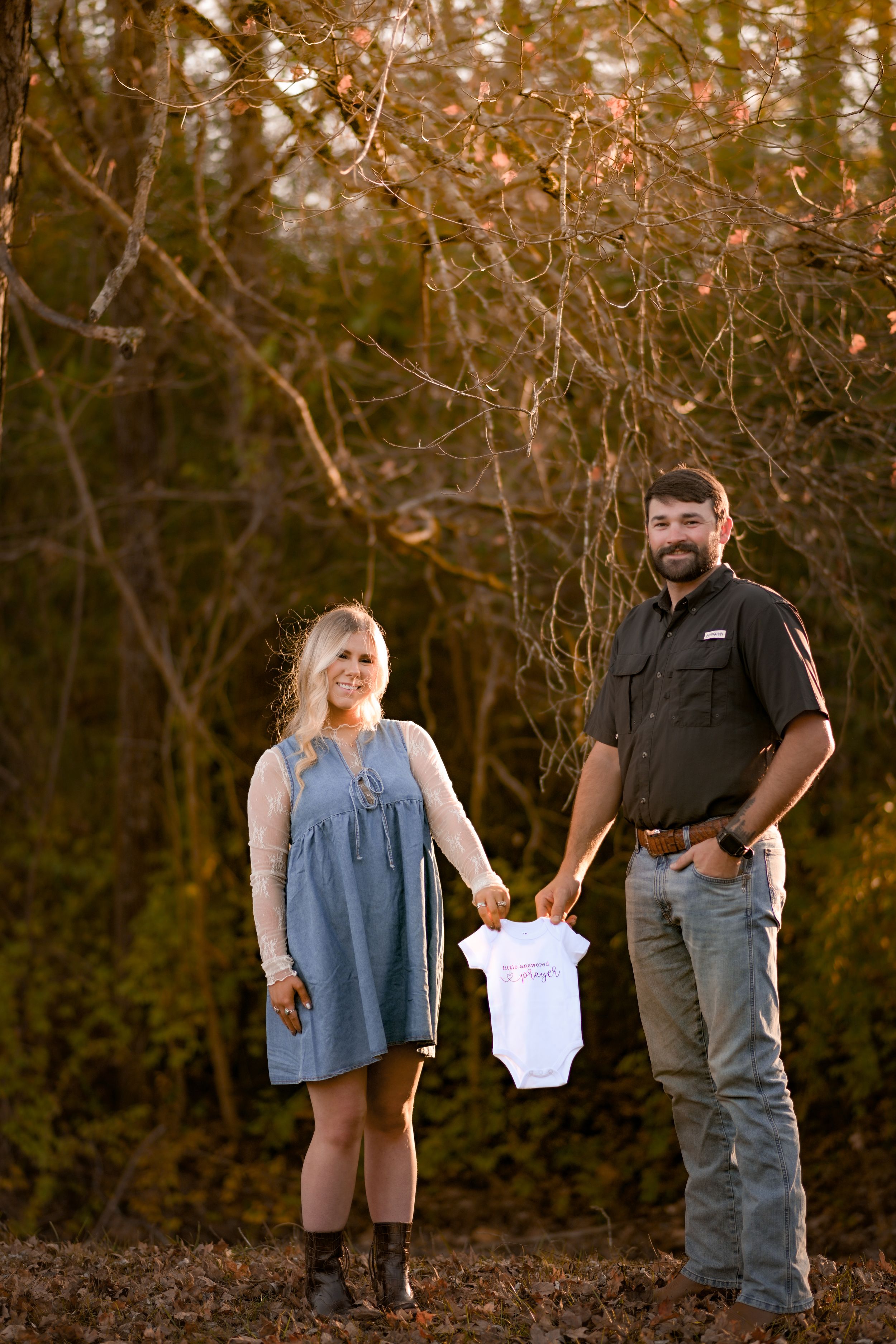 A couple holds a tiny white onesie during an autumn outdoor pregnancy announcement.