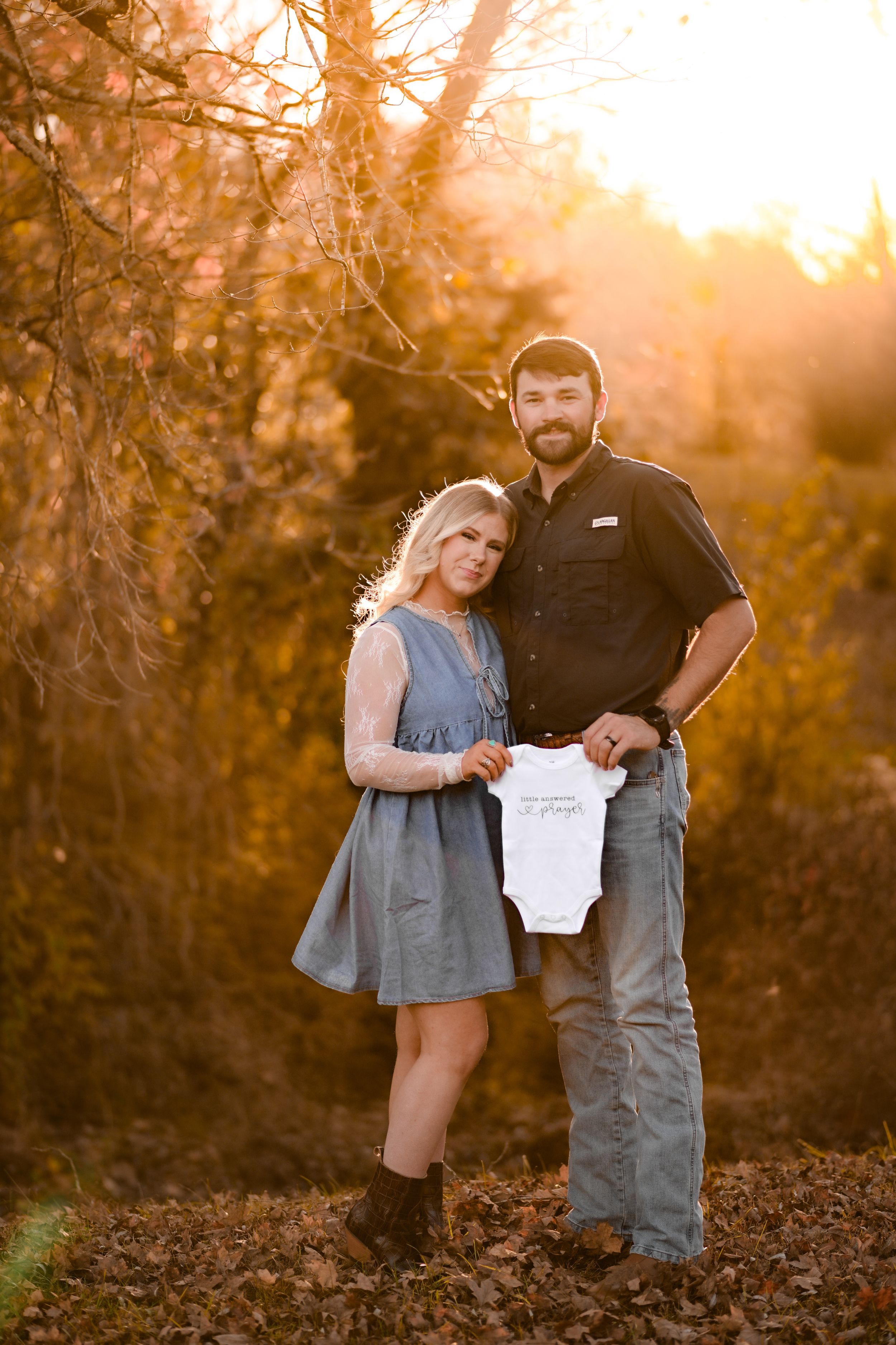 A couple poses with a baby onesie during a sunset pregnancy announcement photoshoot.
