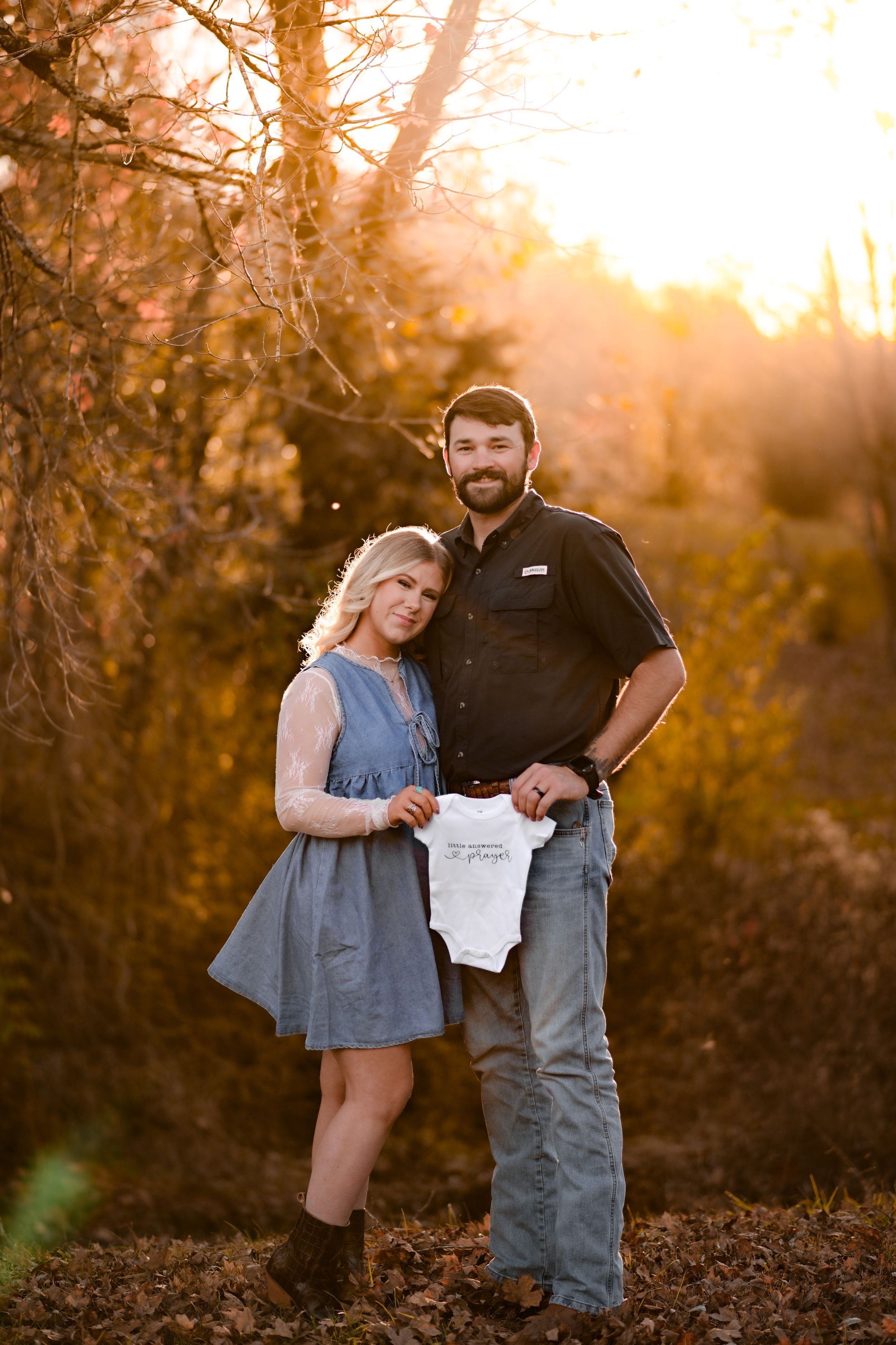 A couple shares their pregnancy news with a white onesie against golden sunset lighting.