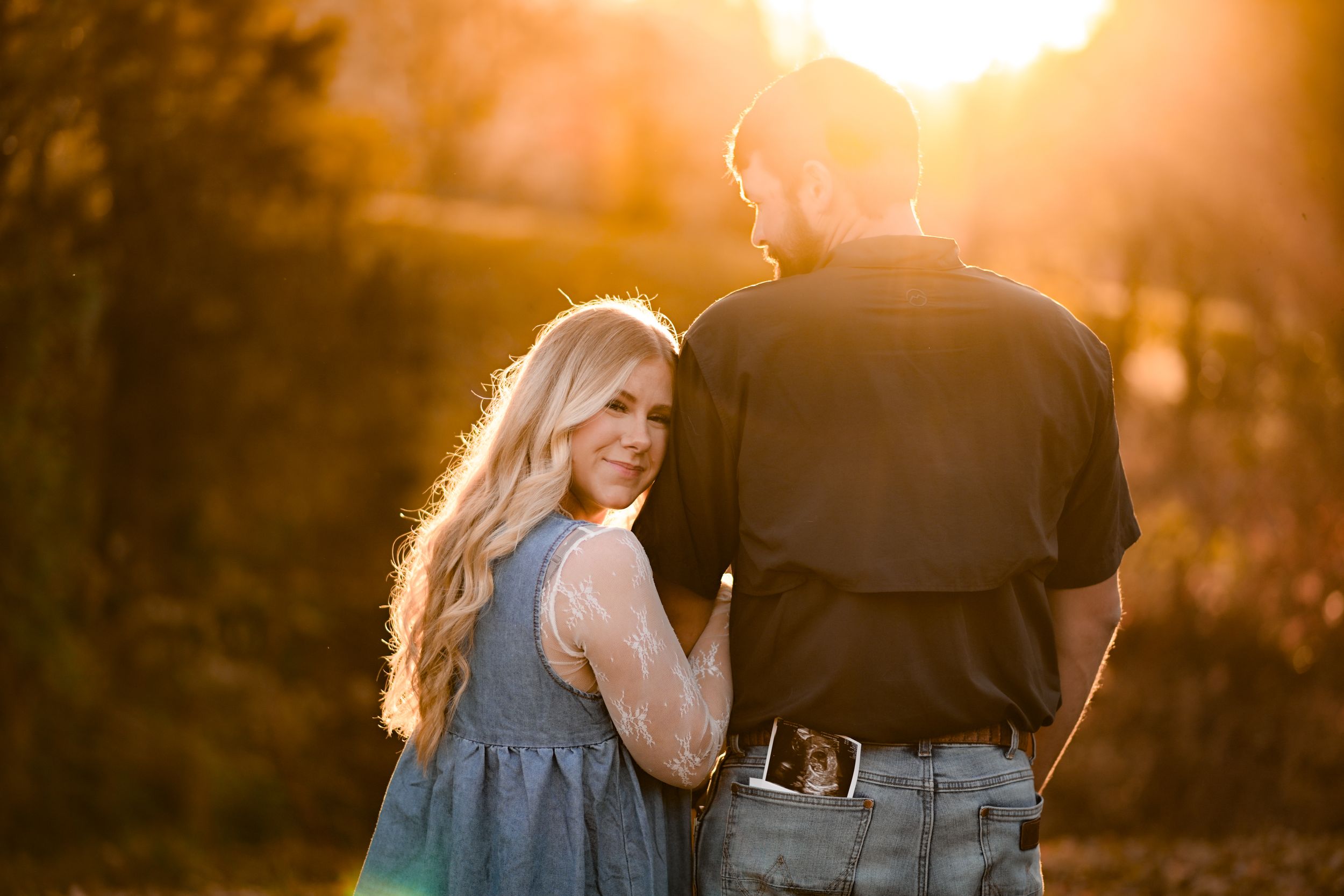 A couple stands together in a golden sunset field with a woman in a blue dress looking back at the camera.