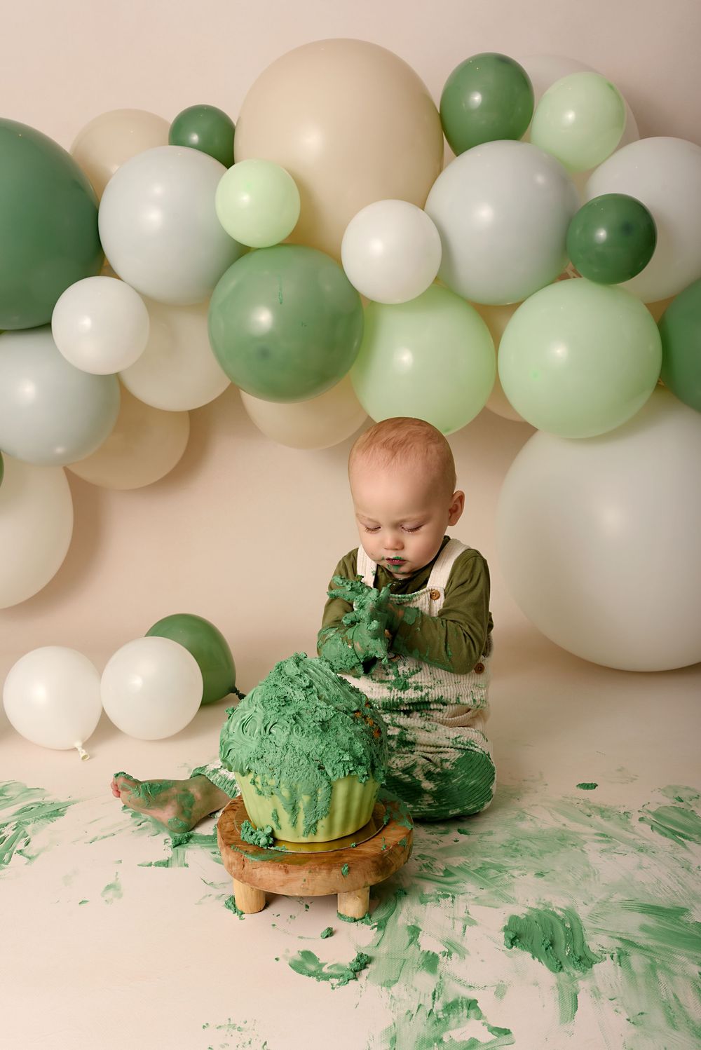 Green and white balloon backdrop frames messy cake smash scene with green frosting scattered on white floor.