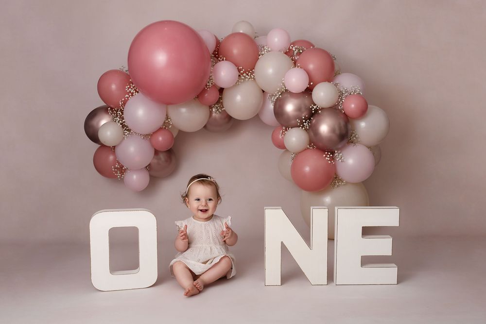 First birthday setup with white letters and pink balloon garland against neutral backdrop.