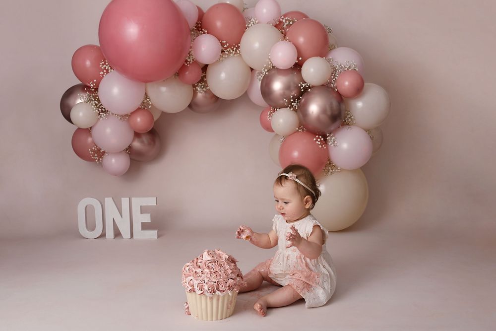 Professional photo setup with pink balloon garland and white ONE letters for birthday celebration.