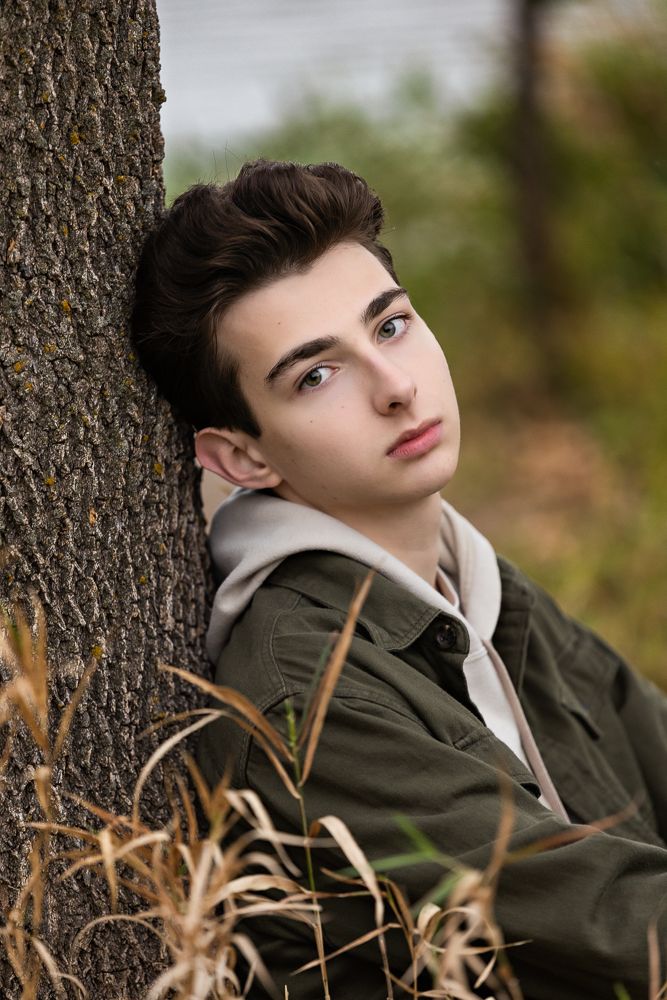 Portrait of someone wearing an olive green jacket and light hoodie leaning against a tree trunk in an outdoor setting.