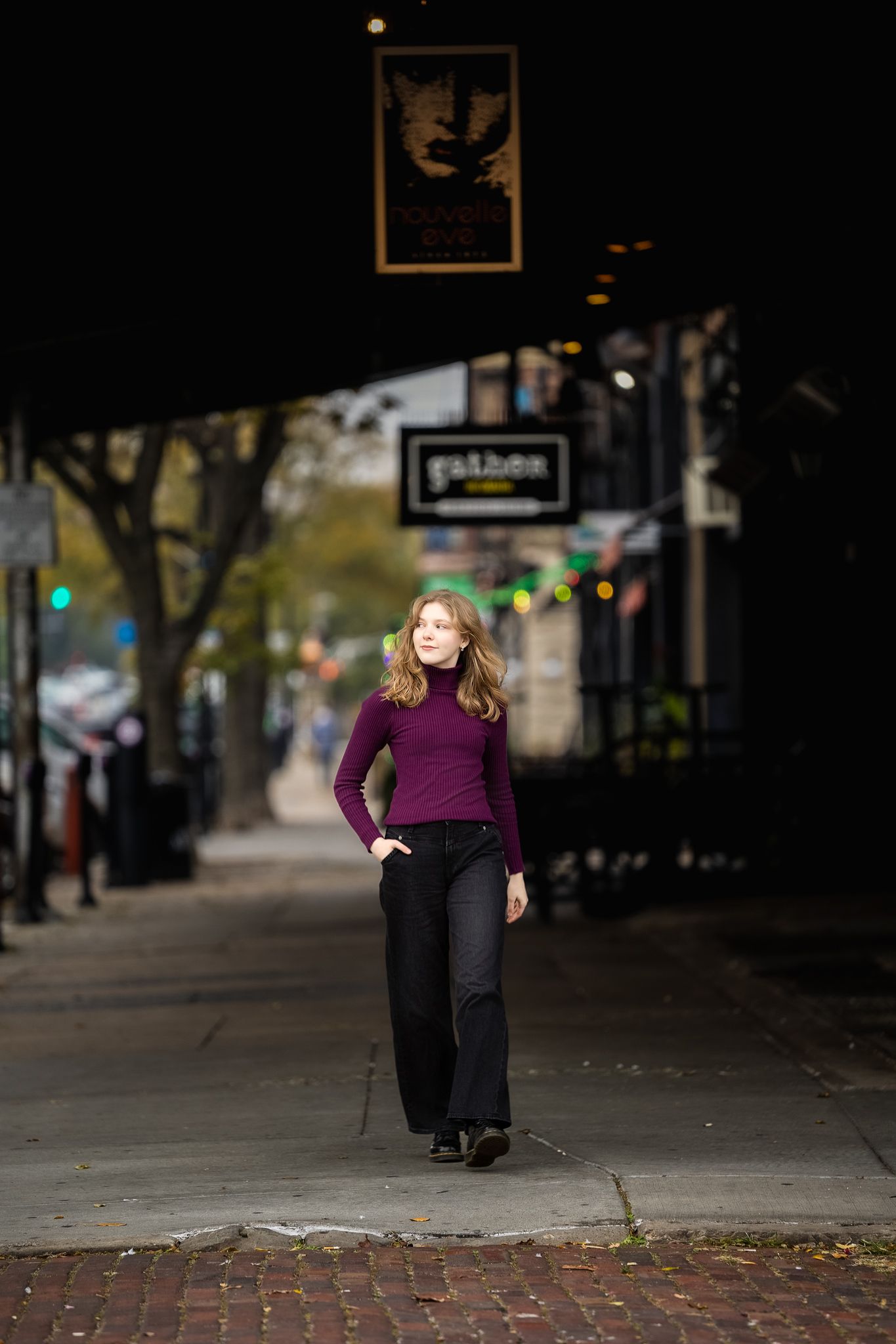 Woman in purple sweater and black pants poses on brick sidewalk in urban setting during daytime.