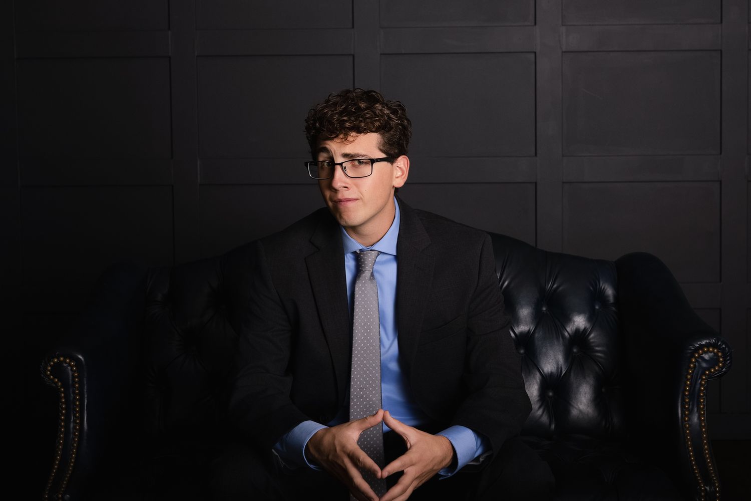 Professional seated in a dark studio wearing a blue dress shirt and glasses while posing in different positions.