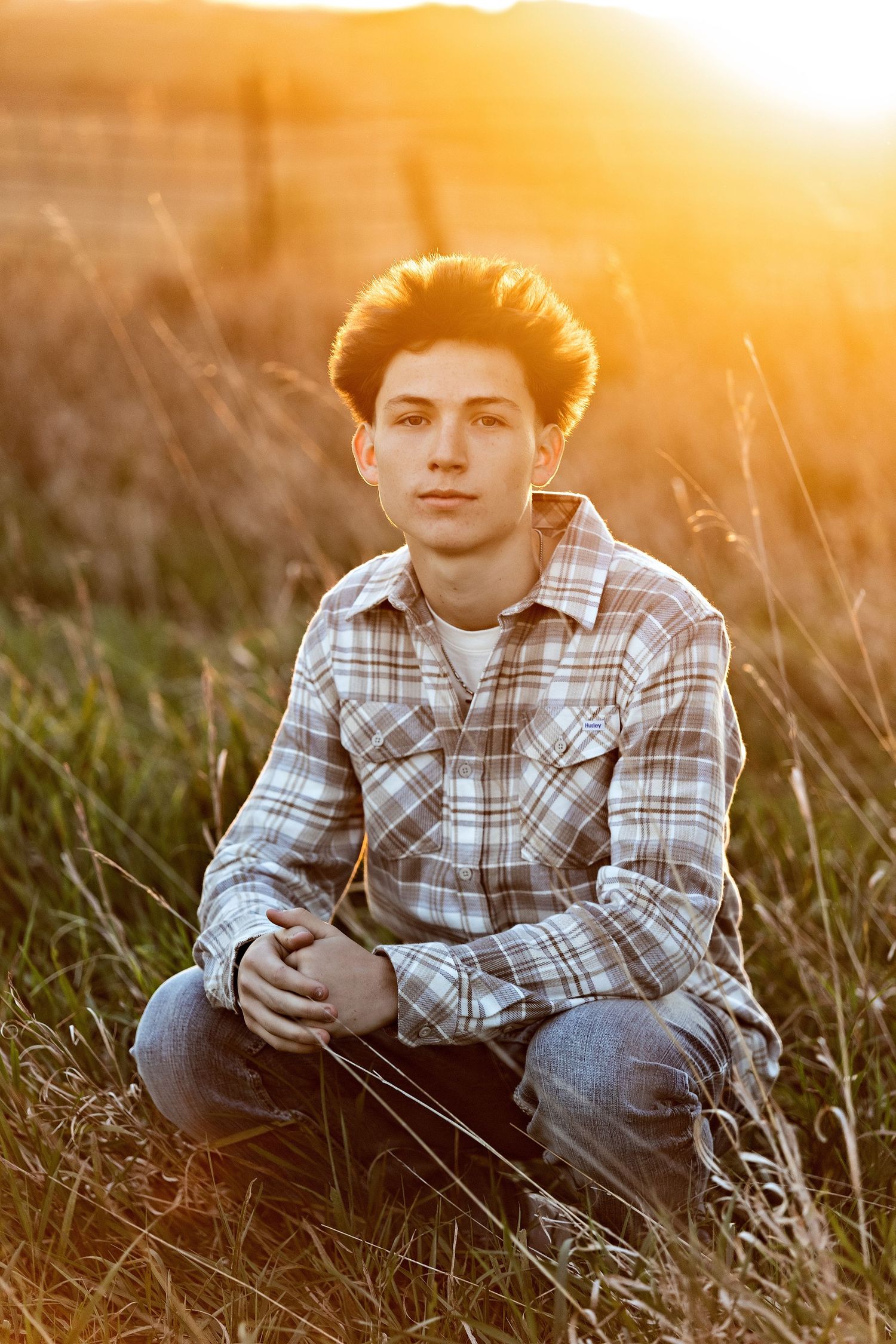 A child in a plaid shirt sits in tall grass at golden hour as sunlight creates a warm backlit glow.