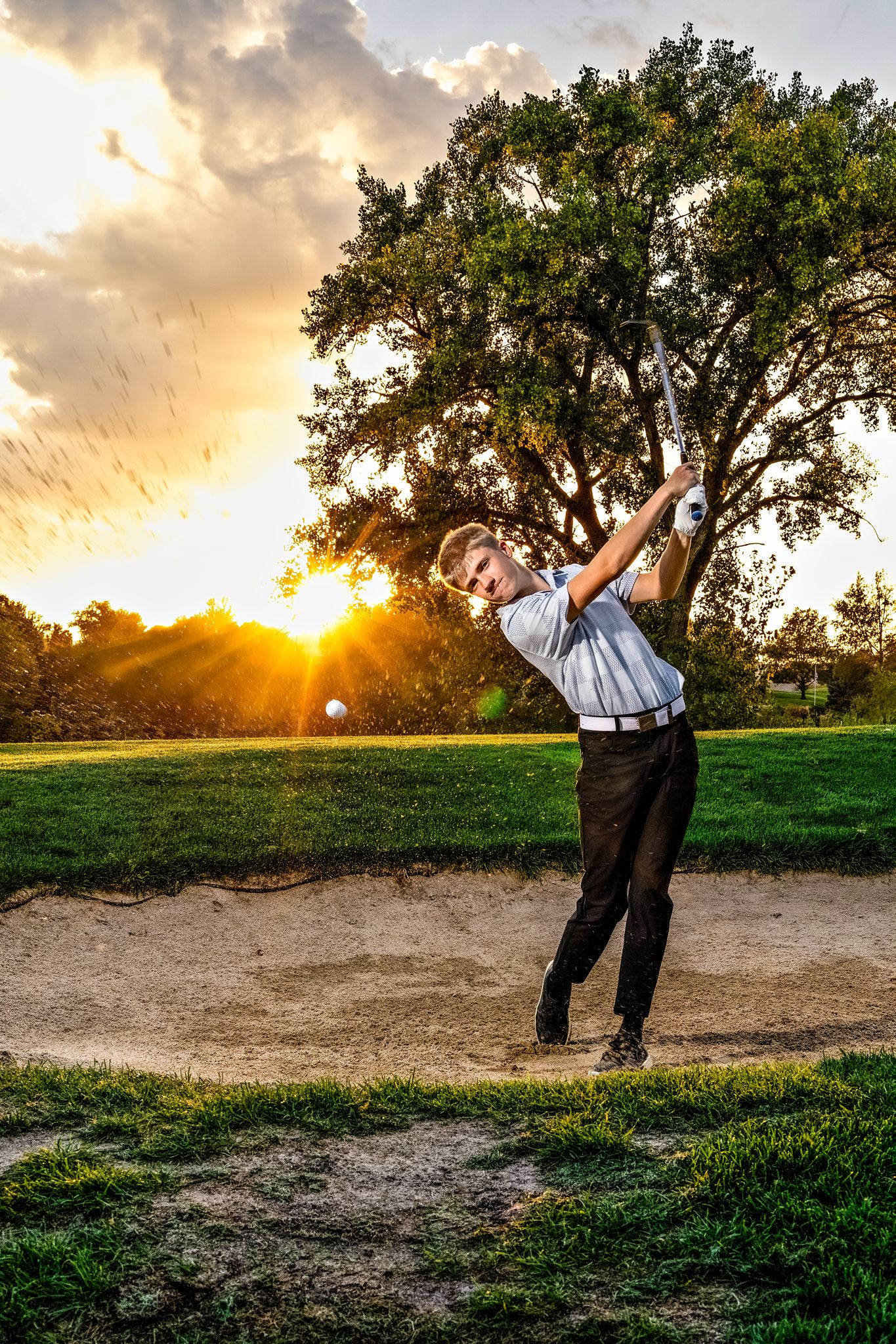 Golfer swings club at sunset on golf course with dramatic sky and trees silhouetted in background.