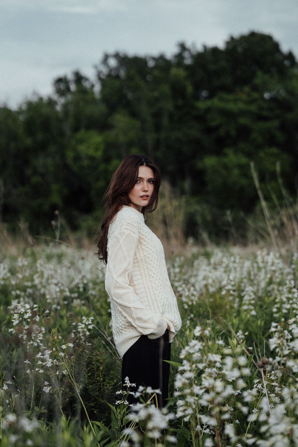Young woman with dark hair in white cable-knit sweater standing among white wildflowers in a lush green field.