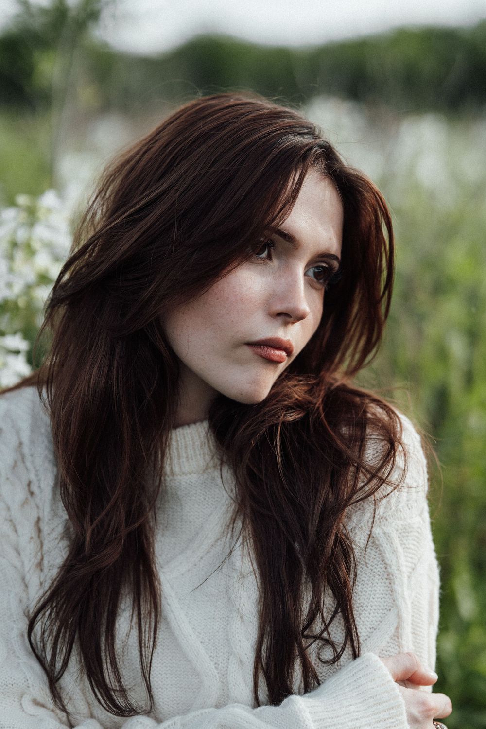 Young woman with long dark hair in white cable-knit sweater gazing thoughtfully in a field of wildflowers.