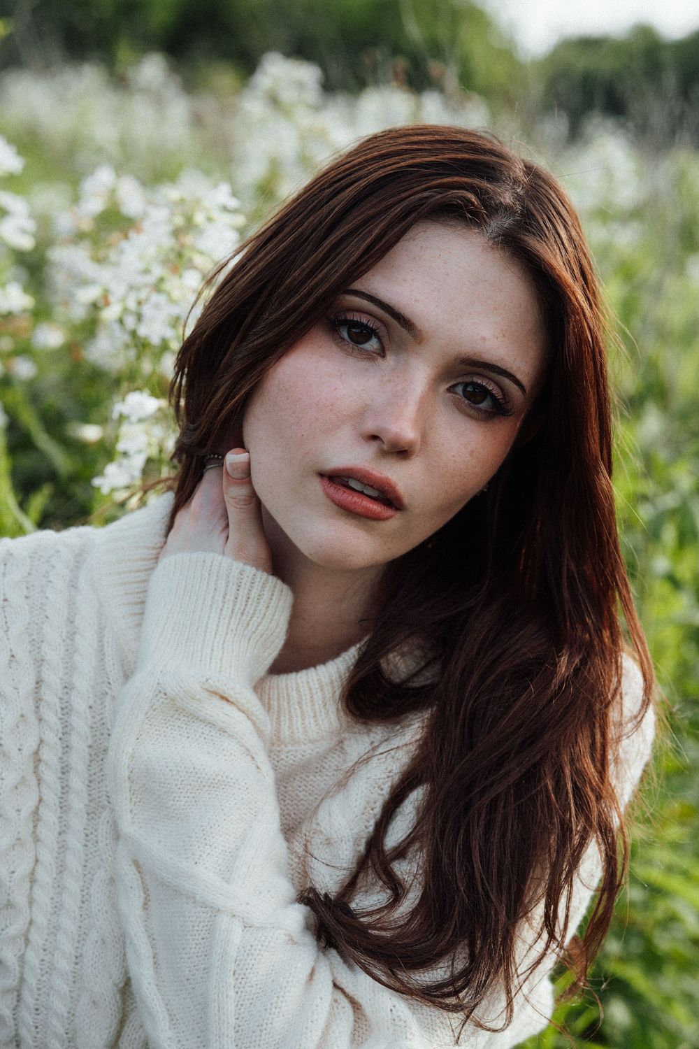 Young woman with red hair and freckles in white knit sweater posing outdoors among wildflowers.