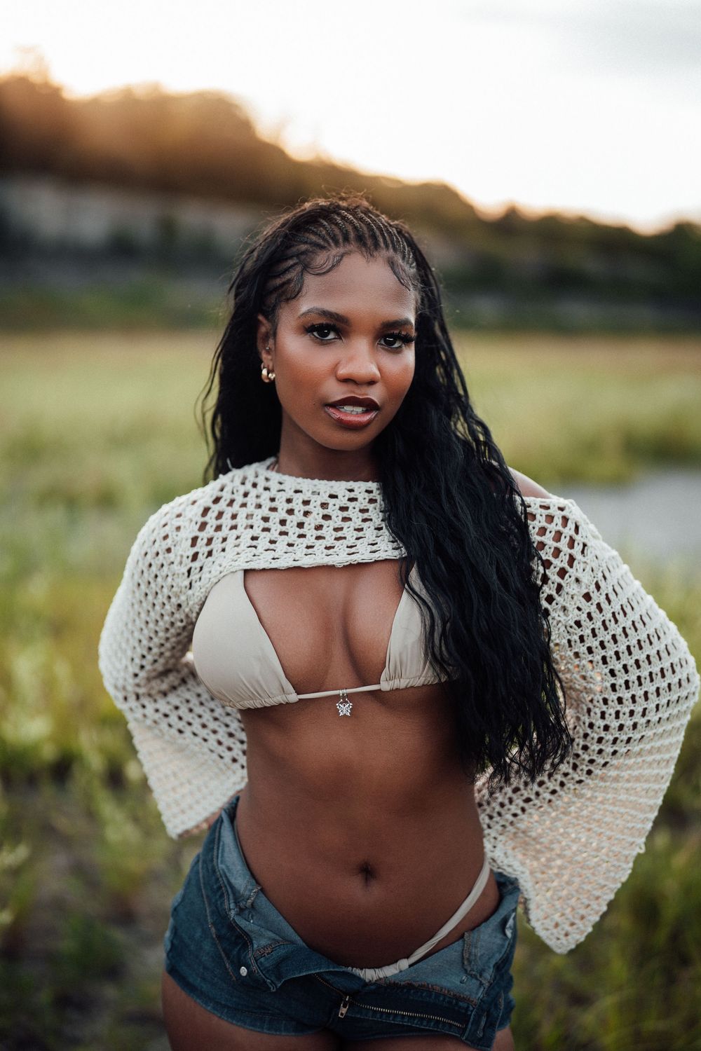Young woman in bikini top, crochet sweater, and denim shorts posing outdoors at golden hour sunset.