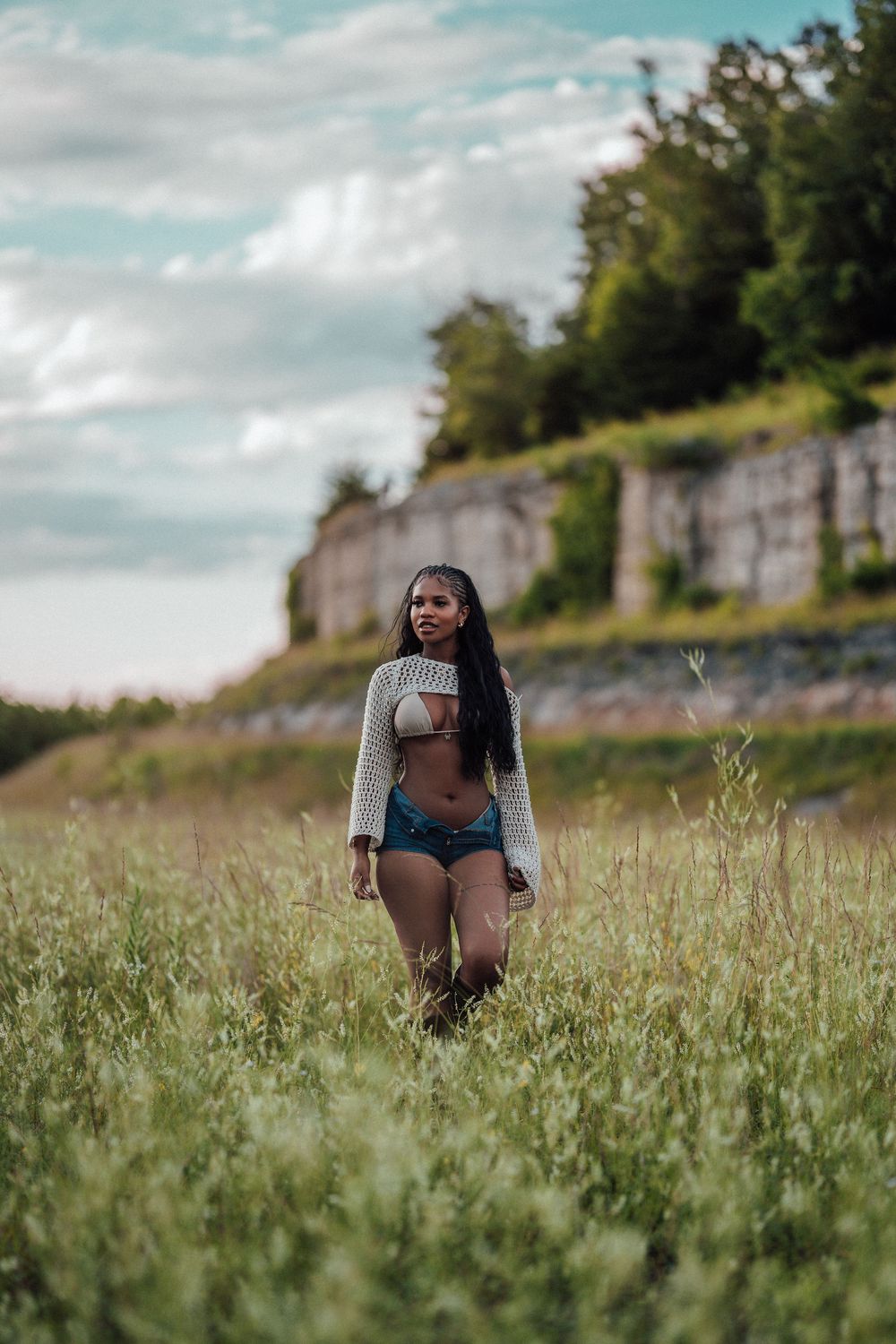 Young woman in crop top and denim shorts walking through tall grass field with stone wall and trees in background.