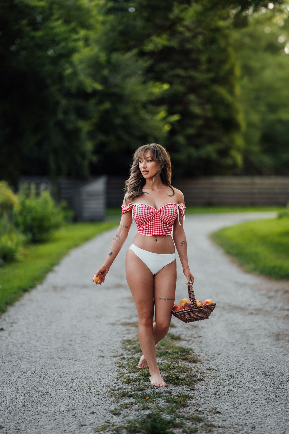 Woman in red checkered bikini top and white bottoms walking barefoot on a gravel path carrying a fruit basket.