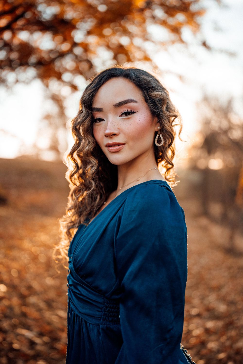 Young woman in teal dress with curly hair poses outdoors during golden autumn sunset.