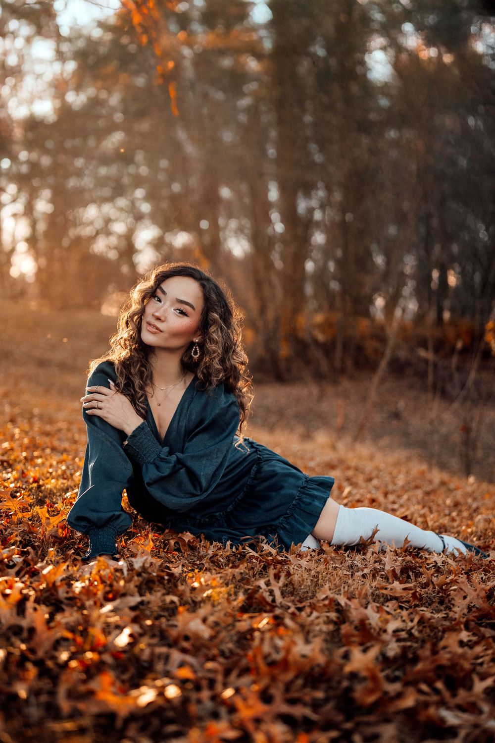 Young woman in teal dress and white knee-high boots sitting on autumn leaves in a golden forest.