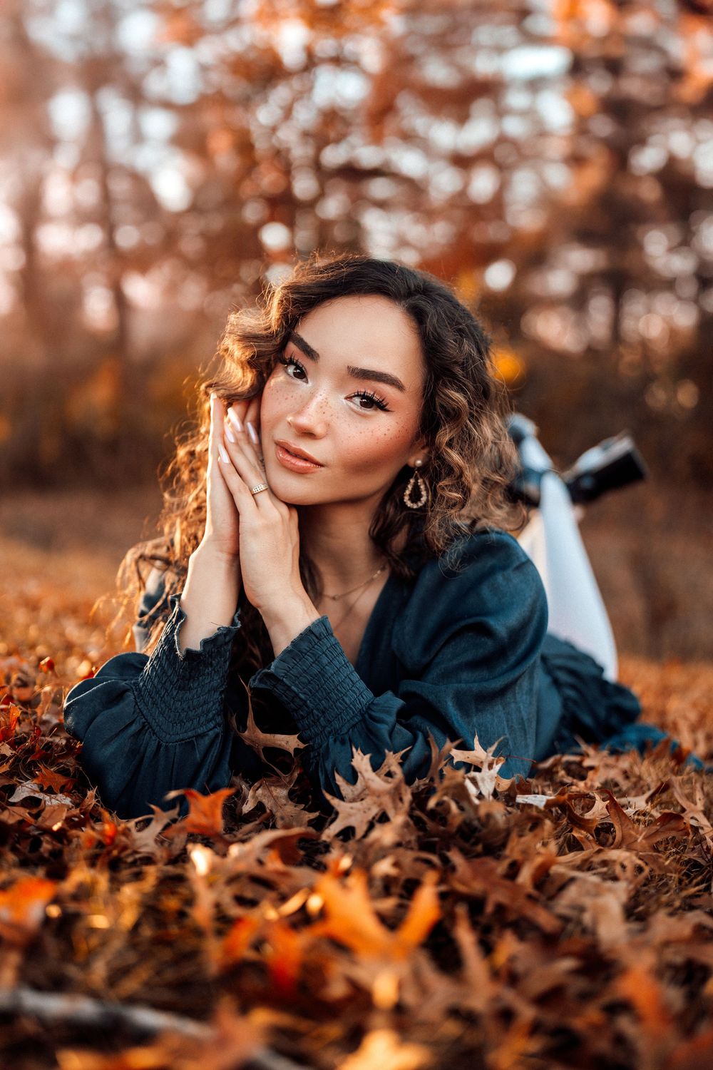 Young woman with curly hair lying in autumn leaves, hands framing face, wearing teal dress in golden fall forest.