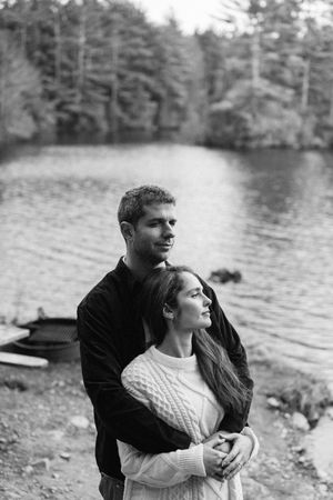 Black and white photo of a couple embracing by a serene lake surrounded by forest trees.