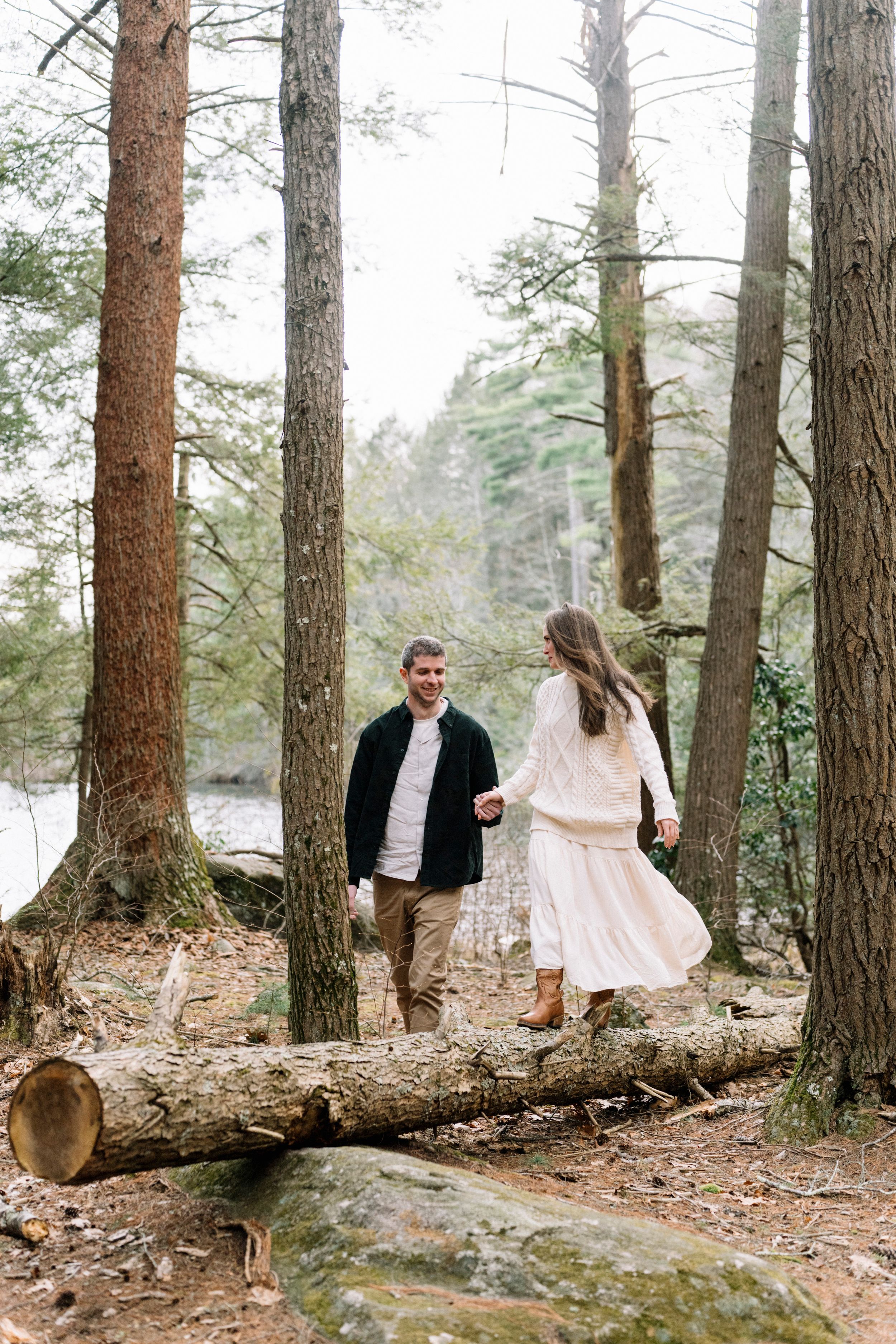 Couple holding hands walking through pine forest over fallen logs near a lake, engagement photo session.
