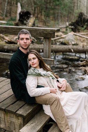 A couple sits together on a wooden bridge by a forest stream, the woman leaning on the man in a white dress.