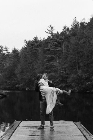 Black and white photo of a man lifting a woman on a dock by a lake, surrounded by forest trees.