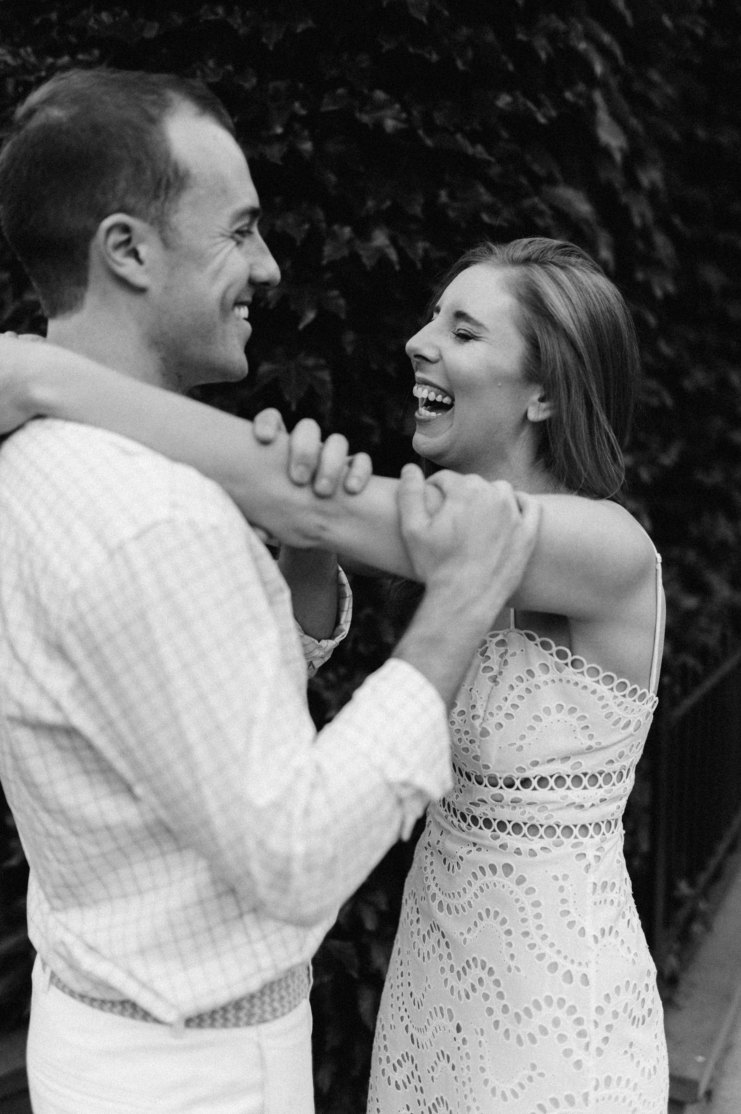 A joyful black and white outdoor photo of a couple in white outfits laughing together during a candid moment.