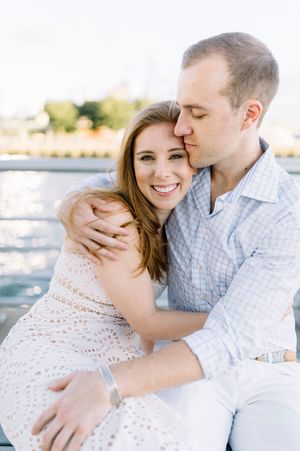 Romantic couple embraces by the water on a sunny day, sharing an intimate moment in light pastel attire.