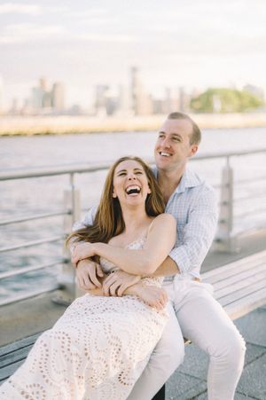 Happy couple laughing together on waterfront bench with city skyline in background on sunny day.
